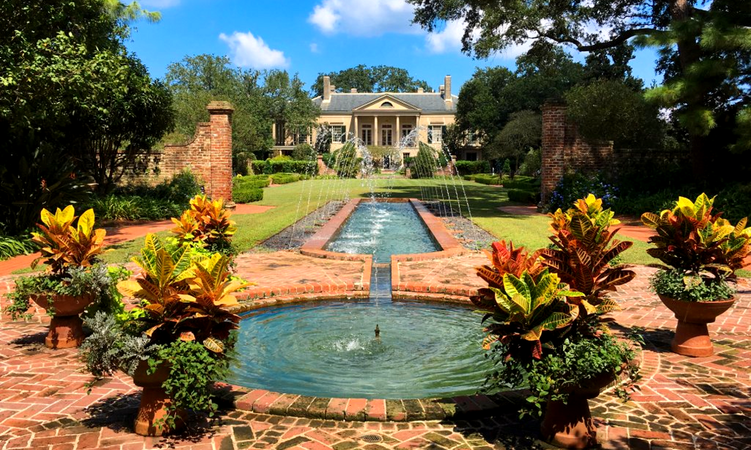 Picture of large house and garden. There is a fountain in the forefront and the white mansion is nestled in the background. 